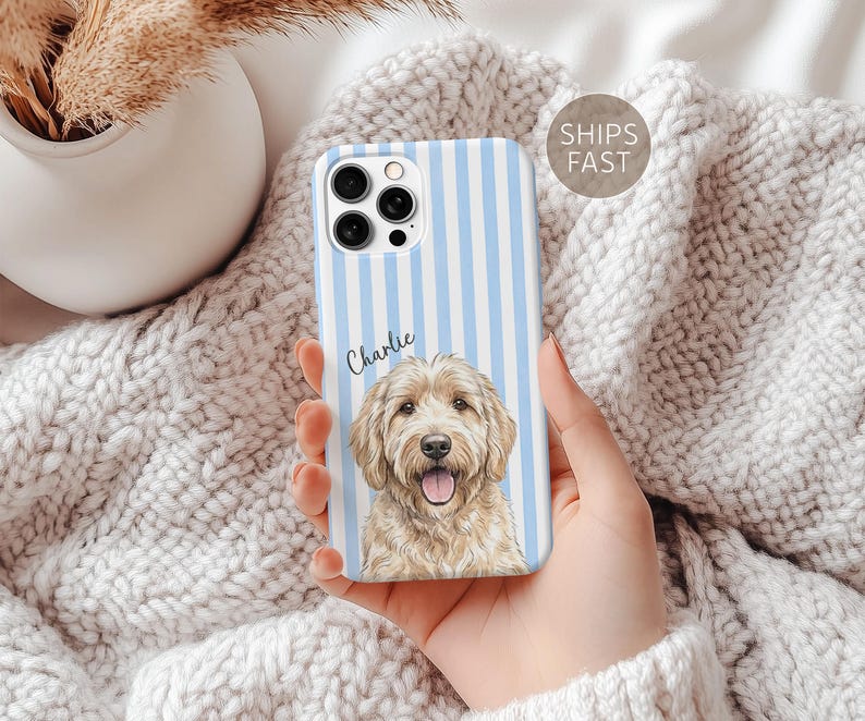 May include: A smartphone case featuring a light blue and white vertical striped pattern. The case displays a detailed illustration of a golden-colored dog with a smiling expression and the name Charlie written in black script above the dog.