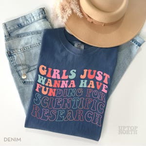 Pode incluir: Uma camiseta azul marinho com o texto "Girls Just Wanna Have Funding For Scientific Research" em cores do arco-&iacute;ris. O texto est&aacute; em um estilo de fonte retr&ocirc;.