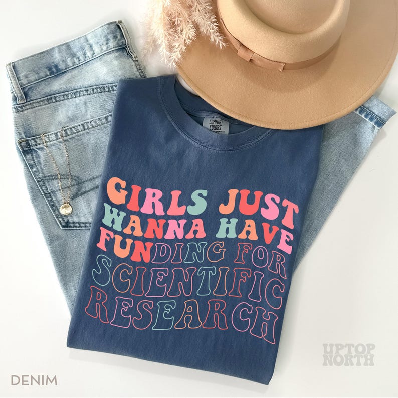 Pode incluir: Uma camiseta azul marinho com o texto "Girls Just Wanna Have Funding For Scientific Research" em cores do arco-&iacute;ris. O texto est&aacute; em um estilo de fonte retr&ocirc;.