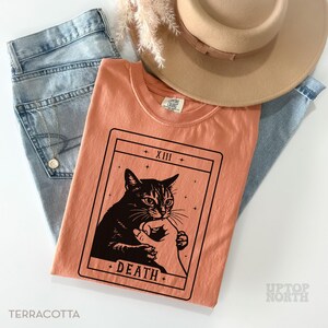 Op de afbeelding: Terracotta T-shirt met een zwarte katten tarotkaart afbeelding met het woord "DEATH". Gecombineerd met een lichtblauwe jeans en een beige hoed. Het label toont "UPTOP NORTH".
