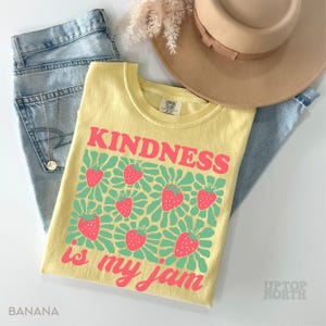 May include: A yellow t-shirt with the text "KINDNESS is my jam" in pink, along with strawberry graphics. The shirt is paired with light blue jeans and a tan hat. The word "BANANA" is printed at the bottom left.