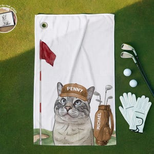 Op de afbeelding: Een witte golfhanddoek met een grafische illustratie van een kat die een bruine pet draagt met de naam Penny erop gedrukt. Naast de kat staat een bruine golftas met het label Michael en een rode vlag aan een vlaggenmast.
