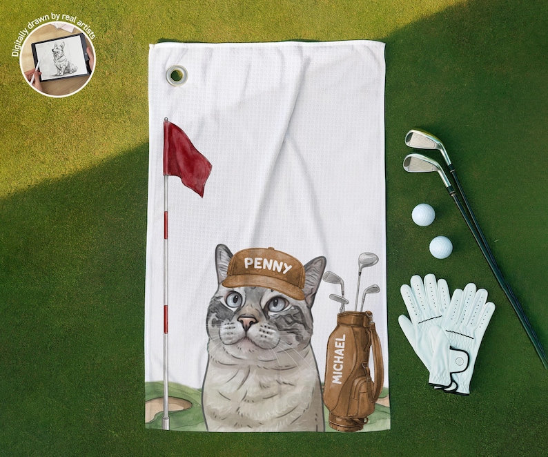 Op de afbeelding: Een witte golfhanddoek met een grafische illustratie van een kat die een bruine pet draagt met de naam Penny erop gedrukt. Naast de kat staat een bruine golftas met het label Michael en een rode vlag aan een vlaggenmast.