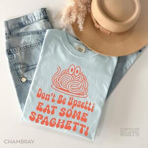 May include: A light blue t-shirt with the text "Don't Be Upsettti EAT SOME SPAGHETTI!" in orange. The shirt is paired with light wash jeans, a tan hat, and a gold necklace.