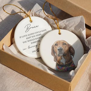May include: Two round white ceramic ornaments with gold hanging strings inside a cardboard box. One ornament features a brown dog portrait wearing a colorful collar. The other displays the text Bruce, If love alone could have kept you here, you would have lived forever, in loving memory, and 2006 with a paw print.
