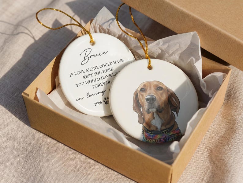 May include: Two round white ceramic ornaments with gold hanging strings inside a cardboard box. One ornament features a brown dog portrait wearing a colorful collar. The other displays the text Bruce, If love alone could have kept you here, you would have lived forever, in loving memory, and 2006 with a paw print.