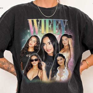 May include: A black crewneck t-shirt featuring a large graphic print with the word WIFEY in a rainbow gradient font. The design includes a collage of five photographs of the same person, set against a nebula-like background with stars.