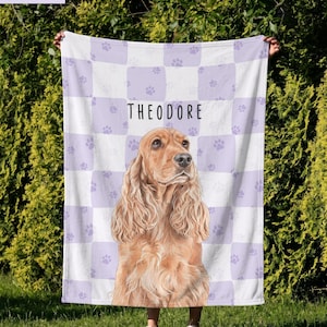 May include: A rectangular fleece blanket featuring a light purple and white checkered pattern with small paw prints. The center displays a realistic illustration of a golden-brown spaniel dog above the name THEODORE in black text.