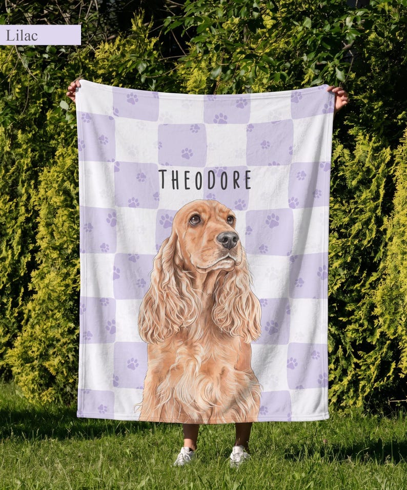 May include: A rectangular fleece blanket featuring a light purple and white checkered pattern with small paw prints. The center displays a realistic illustration of a golden-brown spaniel dog above the name THEODORE in black text.