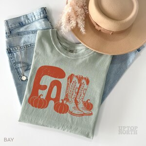 May include: A light green t-shirt with an orange graphic that says "FALL" with pumpkins and cowboy boots. The shirt is paired with light blue jeans, a tan hat, and decorative dried flowers. The word "BAY" is printed on the bottom left.