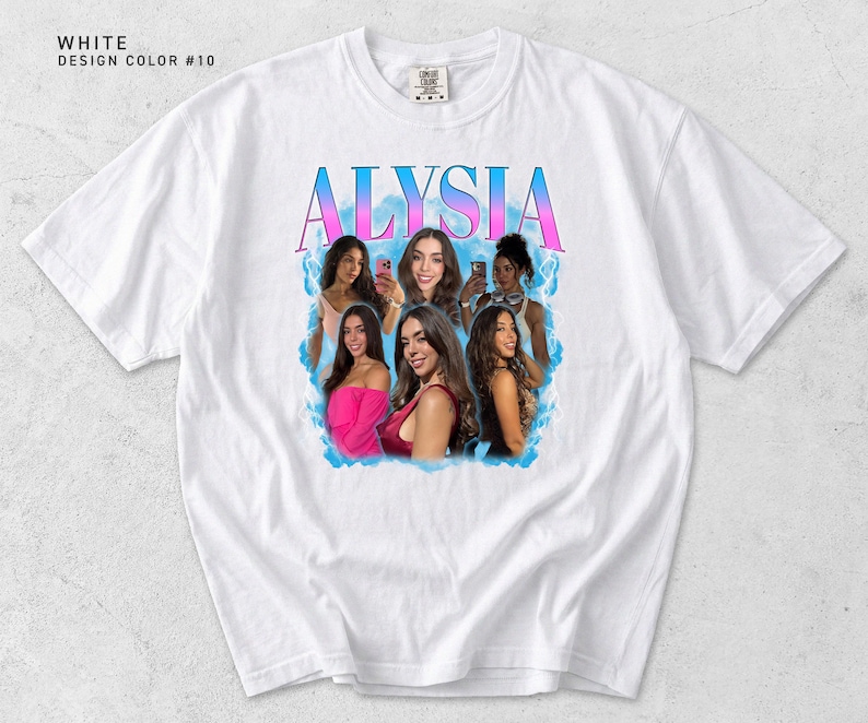 May include: White t-shirt with a graphic design featuring the name "ALYISIA" in a gradient of pink and blue. The design includes images of several women surrounded by a blue cloud-like effect. The shirt has a crew neck and short sleeves.