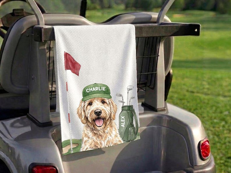 Op de afbeelding: Een witte golfhanddoek met wafelstructuur voorzien van een aquarelillustratie van een goudkleurige hond met een groene pet waarop de naam CHARLIE staat, een rode golfvlag en een groene golfbag met de naam ALEX aan de zijkant gedrukt.