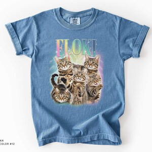 May include: A blue jean colored t-shirt featuring a graphic print of five brown tabby cats arranged in a cluster. Above the cats, the name FLOKI is printed in a rainbow gradient font. The design includes a soft, glowing light effect behind the cats.