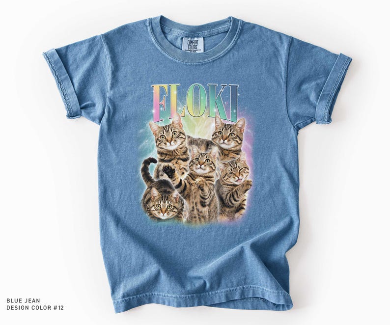 May include: A blue jean colored t-shirt featuring a graphic print of five brown tabby cats arranged in a cluster. Above the cats, the name FLOKI is printed in a rainbow gradient font. The design includes a soft, glowing light effect behind the cats.