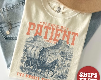 May include: Ivory-colored t-shirt with the text "PLEASE BE PATIENT WITH ME I'M FROM THE 1900S" in coral. The graphic depicts a covered wagon and horse in a desert landscape. A tan hat and blue jeans are in the background.