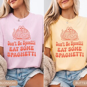 May include: Two t-shirts, one pink and one yellow, each with a spaghetti graphic and the text "Don't Be Upsettti EAT SOME SPAGHETTI!". The shirts are short-sleeved and worn with denim shorts. The pink shirt is labeled "Blossom" and the yellow shirt is labeled "Butter".