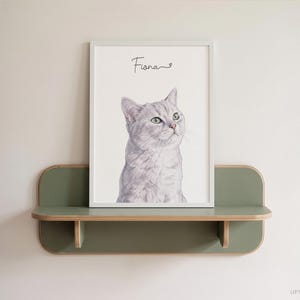 Op de afbeelding: Een ingelijste portretafdruk van een lichtgrijze kat met groene ogen, geplaatst op een wandplank in saliegroen. De naam Fiona staat in zwarte cursieve letters boven de illustratie van de kat op een witte achtergrond.