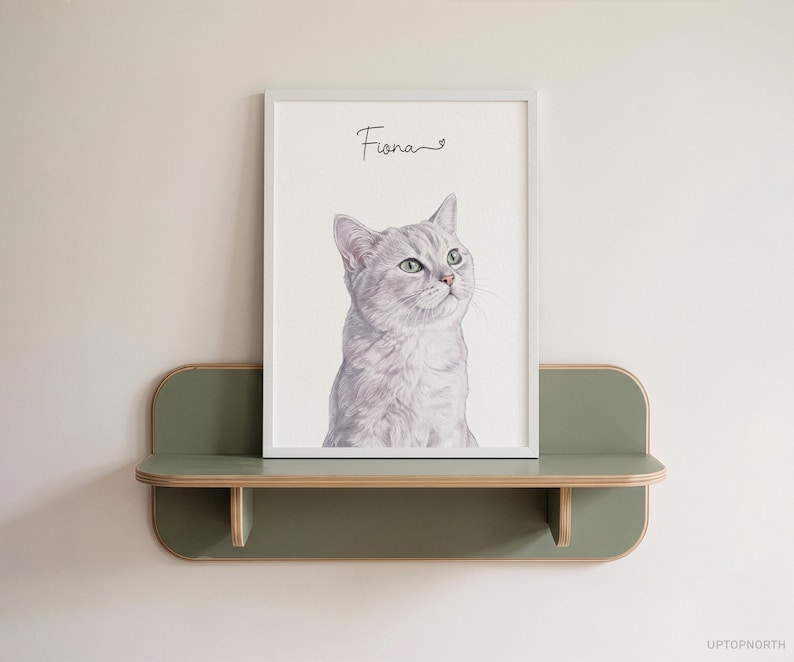 Op de afbeelding: Een ingelijste portretafdruk van een lichtgrijze kat met groene ogen, geplaatst op een wandplank in saliegroen. De naam Fiona staat in zwarte cursieve letters boven de illustratie van de kat op een witte achtergrond.