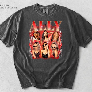 May include: A dark gray t-shirt featuring a graphic of the word "ALLY" in red with a vintage design. The graphic includes images of six women. The shirt has a crew neck and short sleeves.