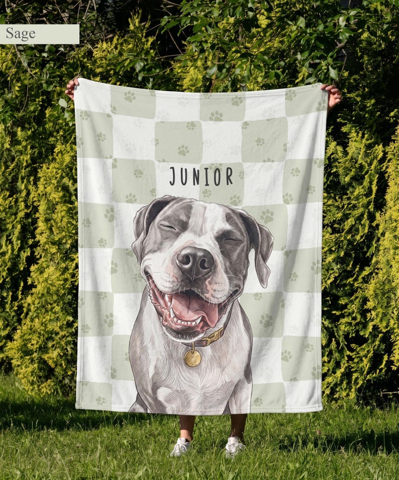 May include: A soft blanket featuring a sage green and white checkered pattern with small paw prints. The center displays a large illustration of a smiling dog with gray and white fur, wearing a collar with a tag, and the name JUNIOR printed above.