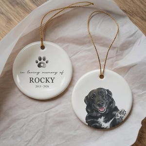 May include: Two round white ceramic ornaments with gold hanging strings. One ornament features a black paw print, the text "in loving memory of ROCKY" and "2013-2026". The other ornament displays a detailed illustration of a black dog with a white chest.
