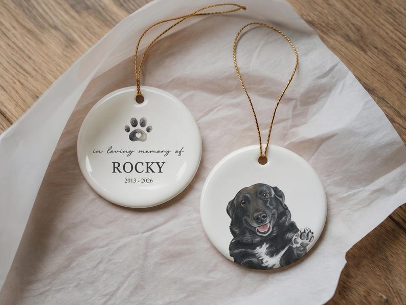 May include: Two round white ceramic ornaments with gold hanging strings. One ornament features a black paw print, the text "in loving memory of ROCKY" and "2013-2026". The other ornament displays a detailed illustration of a black dog with a white chest.