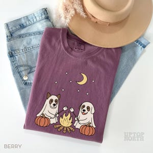 May include: Berry-colored t-shirt featuring a campfire scene with two dogs roasting marshmallows, pumpkins, a crescent moon, and stars. The shirt is paired with light-wash jeans and a tan hat. The text "UPTOP NORTH" is visible.