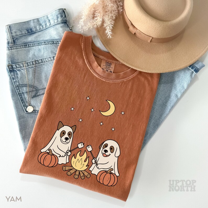 May include: Rust-colored t-shirt featuring a graphic of two dogs roasting marshmallows over a campfire, with pumpkins, stars, and a crescent moon. The shirt is laid on top of light blue jeans, a tan hat, and a necklace.
