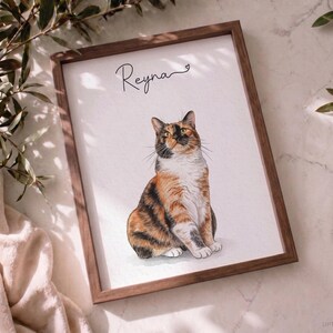 Op de afbeelding: Een ingelijste print met een gedetailleerde illustratie van een zittende lapjeskat. De kat heeft oranje, zwarte en witte vacht. Boven de kat staat de naam Reyna in zwarte cursieve letters met een klein hartje aan het einde.