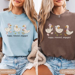 May include: Two t-shirts, one light blue and one brown, each featuring a whimsical illustration of three ducks. The ducks are adorned with accessories and the text "happy, relaxed, engaged" is printed below the ducks.