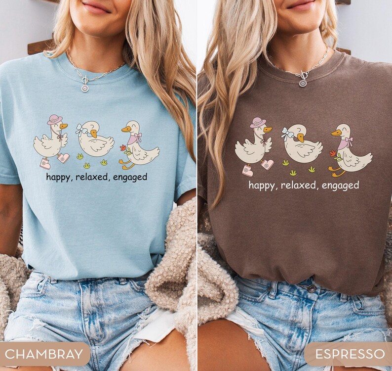 May include: Two t-shirts, one light blue and one brown, each featuring a whimsical illustration of three ducks. The ducks are adorned with accessories and the text "happy, relaxed, engaged" is printed below the ducks.