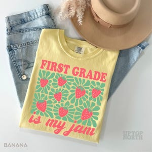 May include: A yellow t-shirt with the text "FIRST GRADE is my jam" in pink, featuring strawberry graphics. The shirt is paired with light blue jeans and a tan hat. The word "BANANA" is printed on the bottom left.