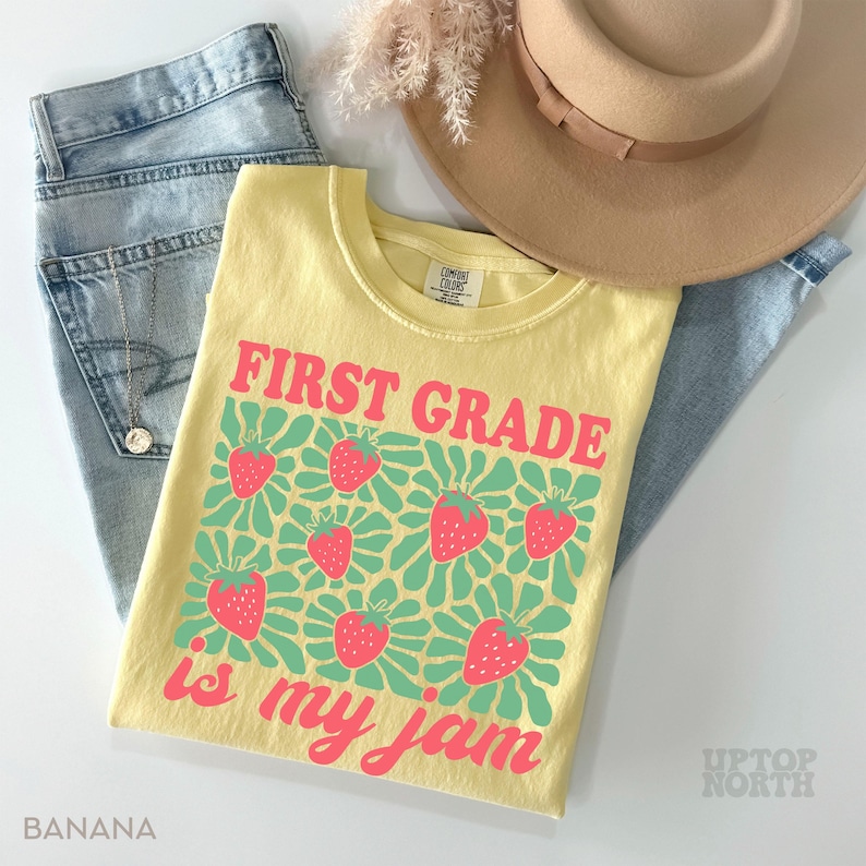 May include: A yellow t-shirt with the text "FIRST GRADE is my jam" in pink, featuring strawberry graphics. The shirt is paired with light blue jeans and a tan hat. The word "BANANA" is printed on the bottom left.