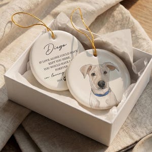 May include: Two round white ceramic ornaments with gold hanging strings. One ornament features a hand-drawn illustration of a light-colored dog with a blue collar. The other displays the text Diego, If love alone could have kept you here, you would have lived forever, in loving memory, and 2014 with a paw print.