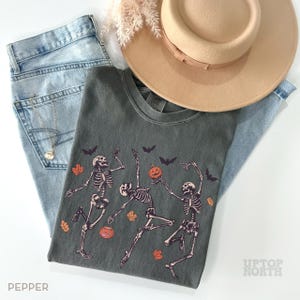 K&ouml;nnte beinhalten: Ein anthrazitfarbenes T-Shirt mit einer Grafik von drei tanzenden Skeletten, orangefarbenen K&uuml;rbissen, schwarzen Flederm&auml;usen und orangefarbenen Herbstbl&auml;ttern. Dazu eine hellblaue Jeans, ein beiger Hut mit breiter Krempe und eine goldene Halskette.
