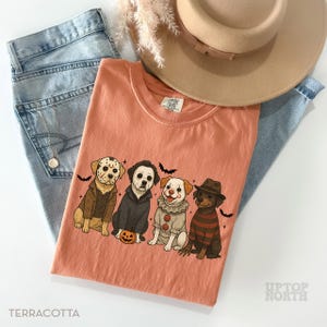 May include: Terracotta-colored t-shirt featuring four cartoon dogs dressed as Halloween characters, including Jason Voorhees, Michael Myers, a clown, and Freddy Krueger. The shirt is paired with light blue jeans and a tan hat. The text "Terracotta" is at the bottom.