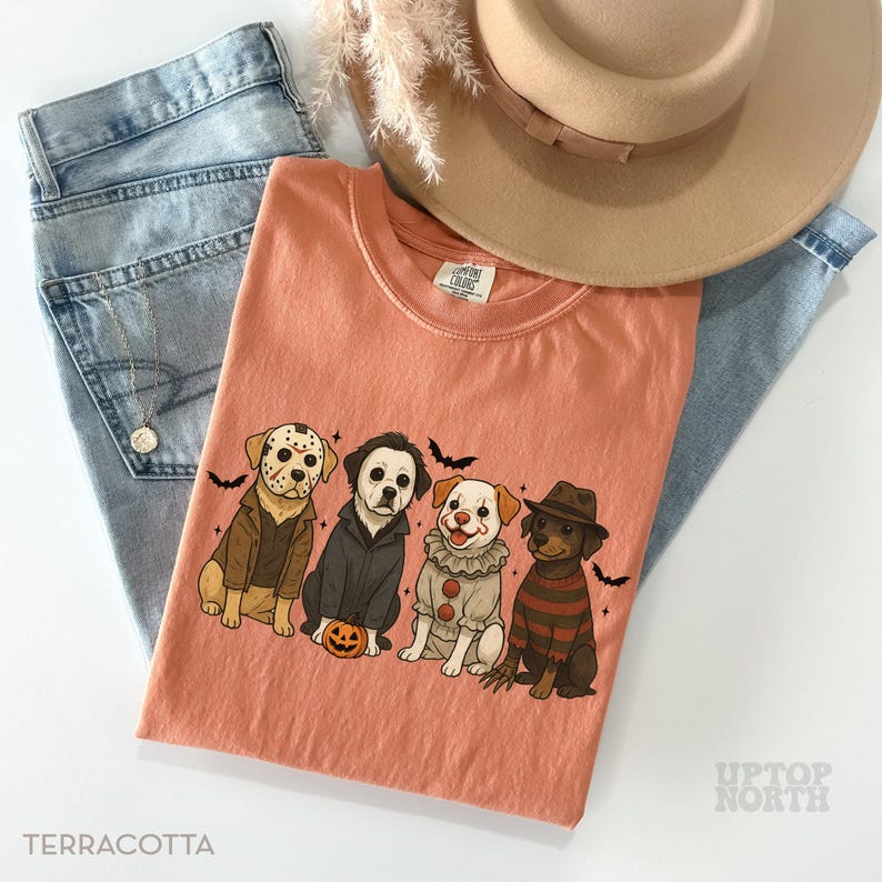 May include: Terracotta-colored t-shirt featuring four cartoon dogs dressed as Halloween characters, including Jason Voorhees, Michael Myers, a clown, and Freddy Krueger. The shirt is paired with light blue jeans and a tan hat. The text "Terracotta" is at the bottom.