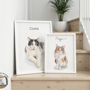 Op de afbeelding: Twee wit ingelijste wandkunstprints met aquarel-stijl portretten van katten. De grotere print toont een zwart-witte kat met het label Cookie, terwijl de kleinere print een lapjeskat toont met het label Maggie in cursief geschreven tekst.