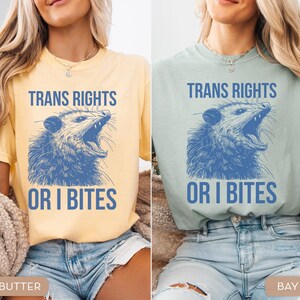 Puede incluir: Dos camisetas, una amarilla mantequilla y otra verde mar, cada una con el texto "TRANS RIGHTS" sobre una ilustraci&oacute;n azul de una zarig&uuml;eya, y las palabras "OR I BITES" debajo.