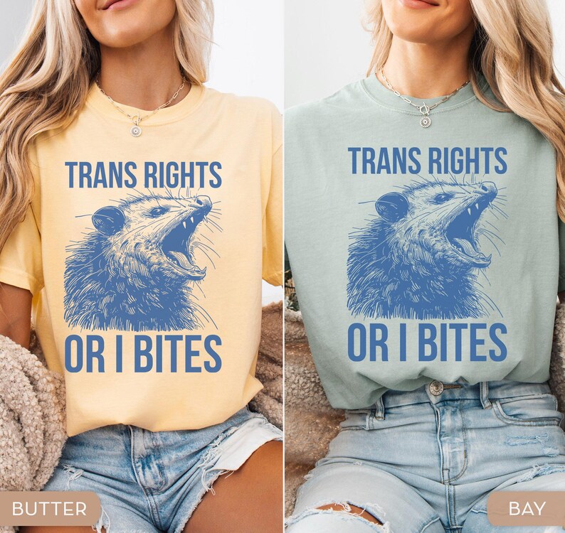 Puede incluir: Dos camisetas, una amarilla mantequilla y otra verde mar, cada una con el texto "TRANS RIGHTS" sobre una ilustraci&oacute;n azul de una zarig&uuml;eya, y las palabras "OR I BITES" debajo.