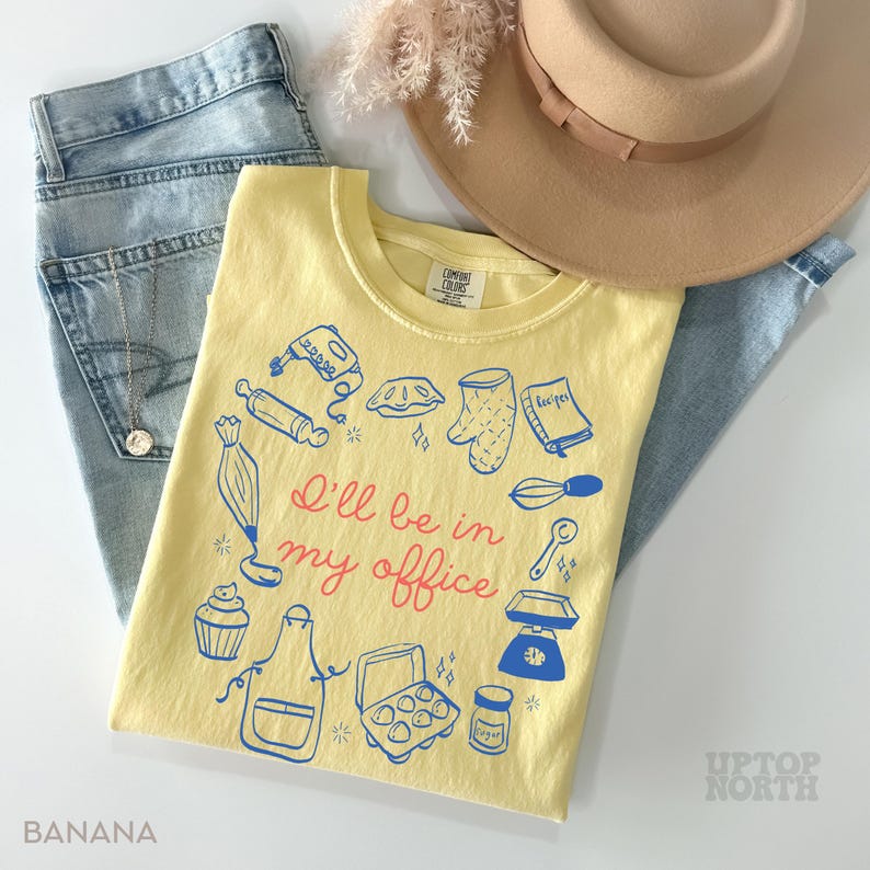 Puede incluir: Camiseta amarilla clara con el texto "I'll be in my office" e ilustraciones azules de utensilios de cocina. La camiseta est&aacute; doblada junto a unos vaqueros azules y un sombrero beige. La palabra "BANANA" est&aacute; impresa abajo a la izquierda.