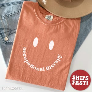 May include: Terracotta-colored t-shirt with a smiley face design. The design includes two white oval eyes and the words "occupational therapy" in a curved line. The shirt is folded and placed on top of blue jeans and a tan hat.