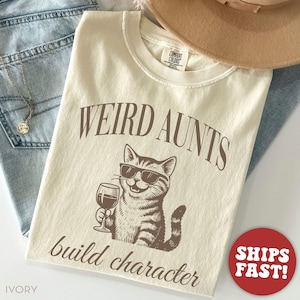 May include: Ivory-colored t-shirt with the text "WEIRD AUNTS build character" and a graphic of a cat wearing sunglasses and holding a wine glass. The shirt is made of a soft material and is a Comfort Colors brand.