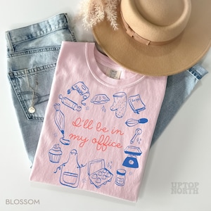 Puede incluir: Una camiseta rosa claro con un gr&aacute;fico azul y rojo de utensilios de reposter&iacute;a y el texto "I'll be in my office". La camiseta se combina con vaqueros azul claro y un sombrero beige. La palabra "Blossom" est&aacute; impresa en la parte inferior izquierda.