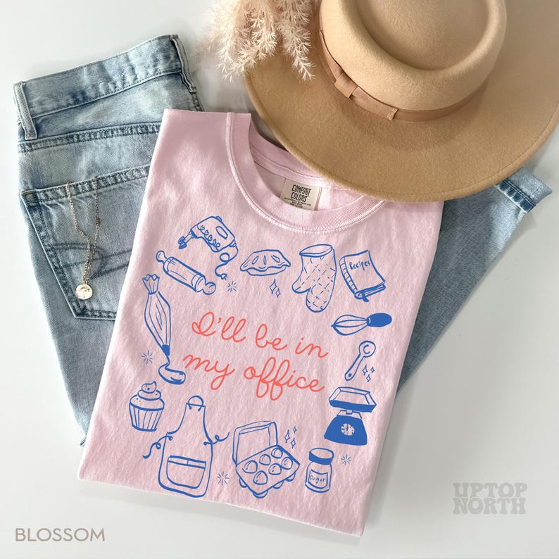 Puede incluir: Una camiseta rosa claro con un gr&aacute;fico azul y rojo de utensilios de reposter&iacute;a y el texto "I'll be in my office". La camiseta se combina con vaqueros azul claro y un sombrero beige. La palabra "Blossom" est&aacute; impresa en la parte inferior izquierda.