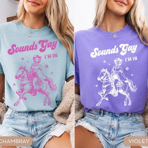 K&ouml;nnte beinhalten: Zwei Frauen tragen T-Shirts mit dem Text "Sounds Gay I'm In" und einer Grafik einer Cowgirl, die ein Pferd reitet. Das T-Shirt links ist hellblau und das T-Shirt rechts ist lila.