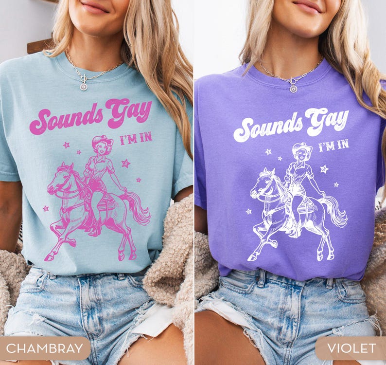K&ouml;nnte beinhalten: Zwei Frauen tragen T-Shirts mit dem Text "Sounds Gay I'm In" und einer Grafik einer Cowgirl, die ein Pferd reitet. Das T-Shirt links ist hellblau und das T-Shirt rechts ist lila.