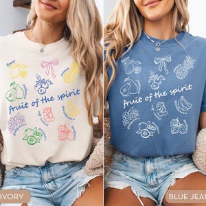May include: Two t-shirts, one ivory and one blue, each featuring a colorful graphic design. The design includes various fruits and the words "fruit of the spirit." The ivory shirt has a multi-colored design, while the blue shirt has a white design.