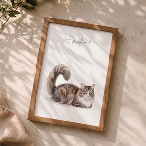 Op de afbeelding: Een ingelijste print met een aquarel-stijl illustratie van een langharige bruin-witte cyperse kat met een pluizige staart. De naam Hazel staat in zwarte cursieve letters boven de kat, vergezeld door een klein hartje.