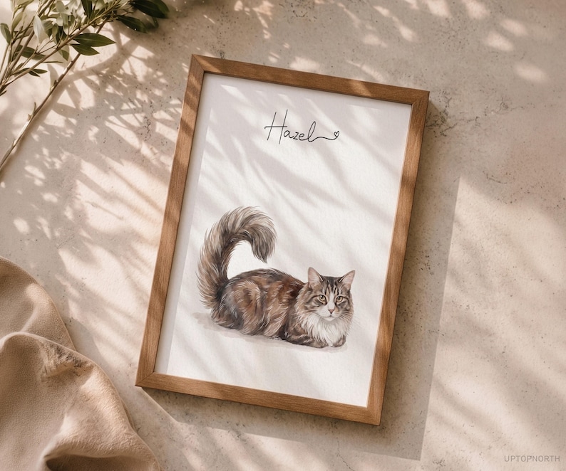 Op de afbeelding: Een ingelijste print met een aquarel-stijl illustratie van een langharige bruin-witte cyperse kat met een pluizige staart. De naam Hazel staat in zwarte cursieve letters boven de kat, vergezeld door een klein hartje.
